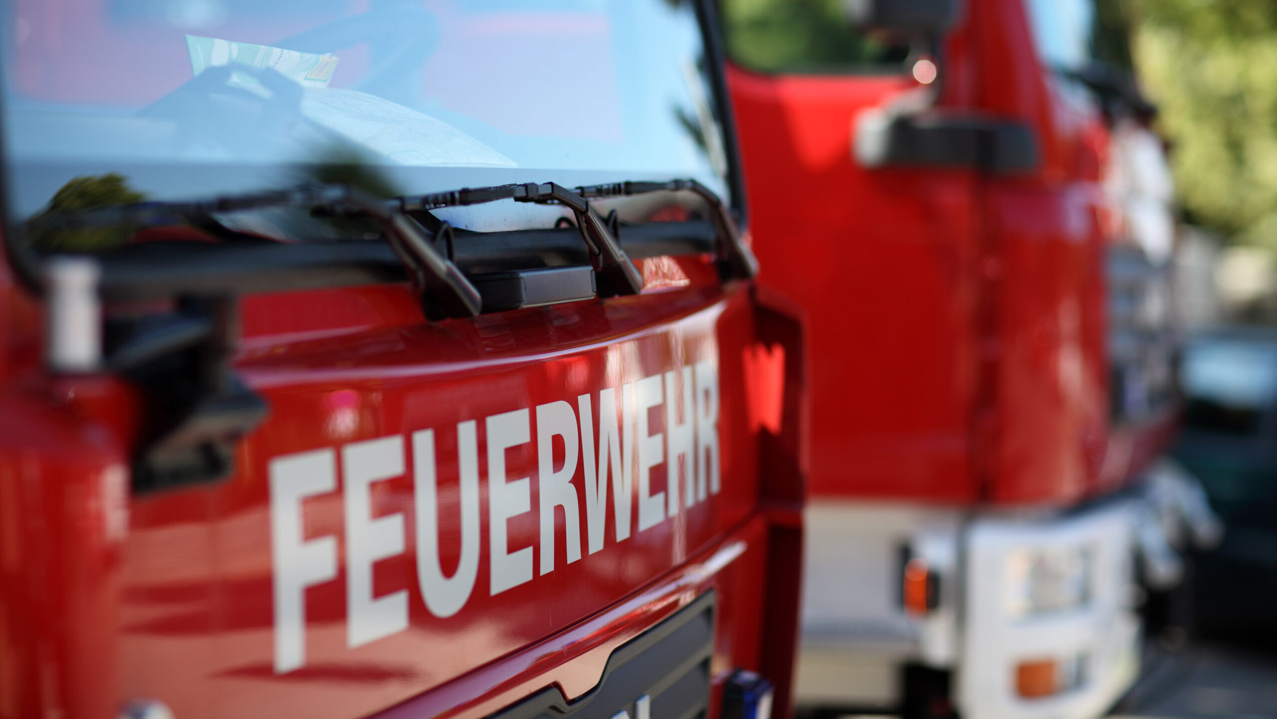 Das Bild zeigt ein rotes Feuerwehrfahrzeug mit dem Schriftzug "FEUERWEHR". Im Hintergrund ist ein weiteres Feuerwehrfahrzeug leicht unscharf zu sehen.